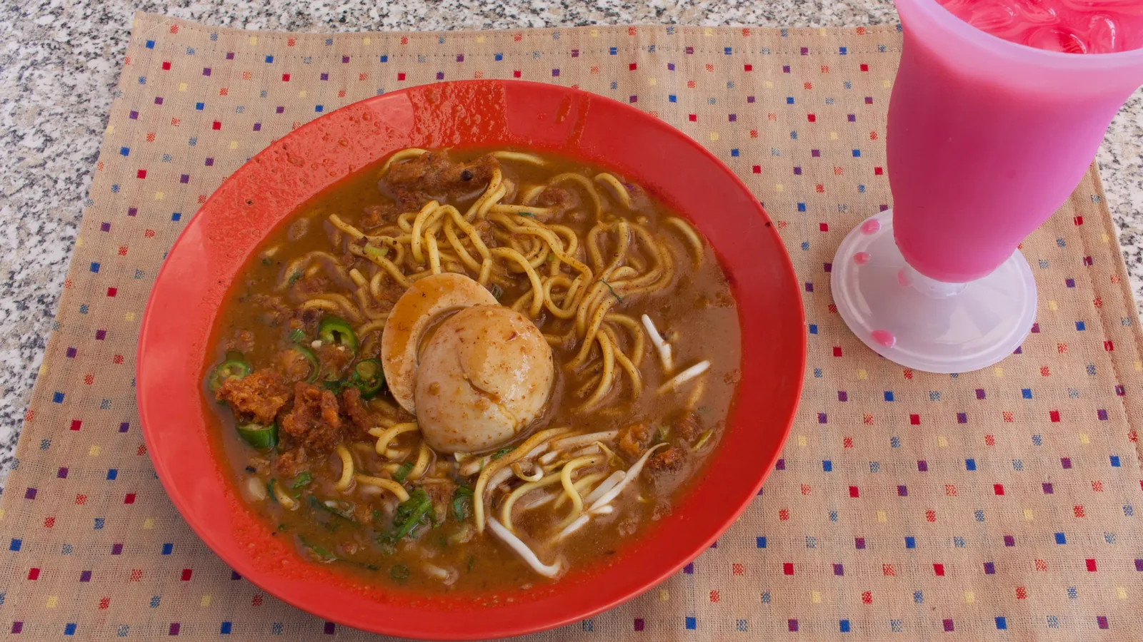 A plate of mee rebus with egg and green chillies beside a glass of pink bandung rose milk drink