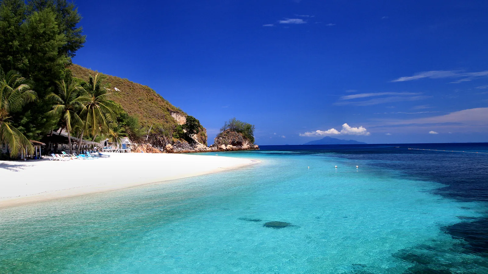 White sand beach and clear turquoise water at Rawa Island off Mersing, Johor, with palm trees and a rocky headland under a deep blue sky