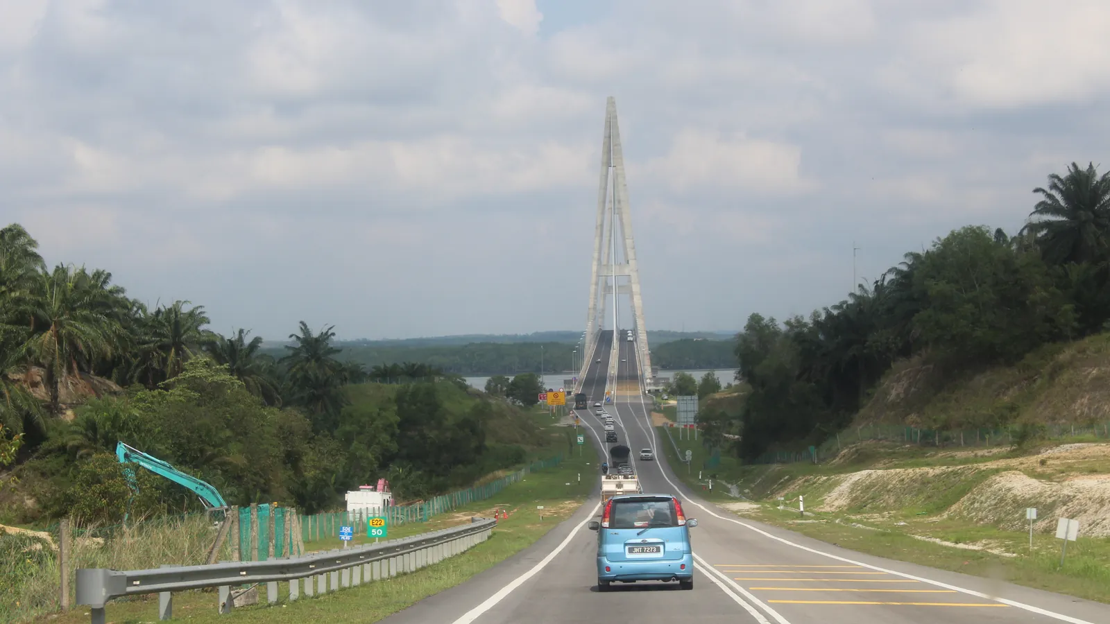 Cars on the Senai–Desaru Expressway heading towards the cable-stayed Sungai Johor Bridge, with oil palms and green hills on both sides