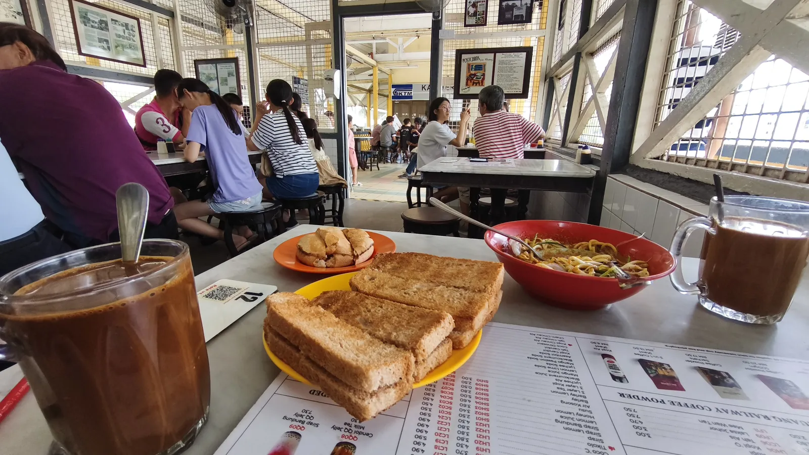 Glasses of kopi, charcoal-toasted kaya bread, a bun and a bowl of noodles on a table inside Kluang Rail Coffee at the old Kluang railway station