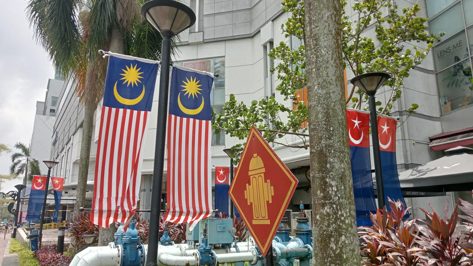 Malaysian Jalur Gemilang flags and blue-and-red Johor state flags hanging from lamp posts outside a mall in Johor Bahru