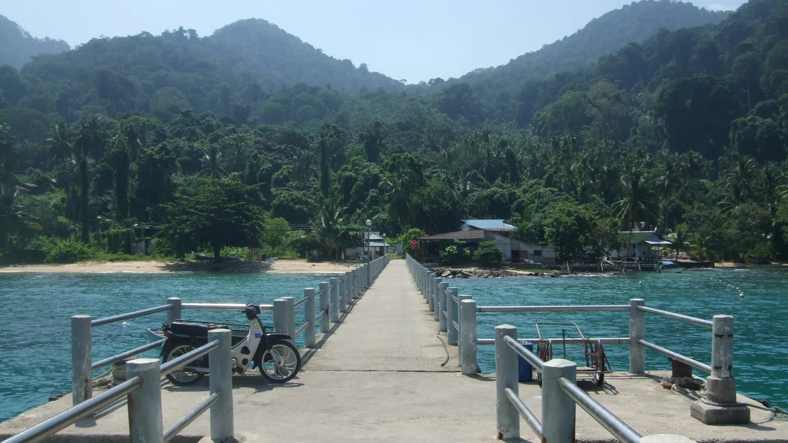 Concrete ferry jetty at Air Batang (ABC) on Tioman Island, leading to a beach and green forested hills over clear turquoise water