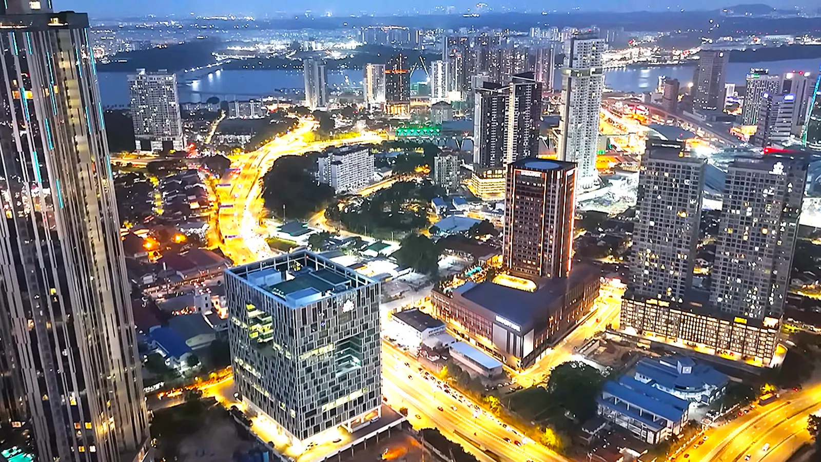 Johor Bahru city centre lit up at night, seen from above, with towers, glowing roads and the Strait of Johor beyond