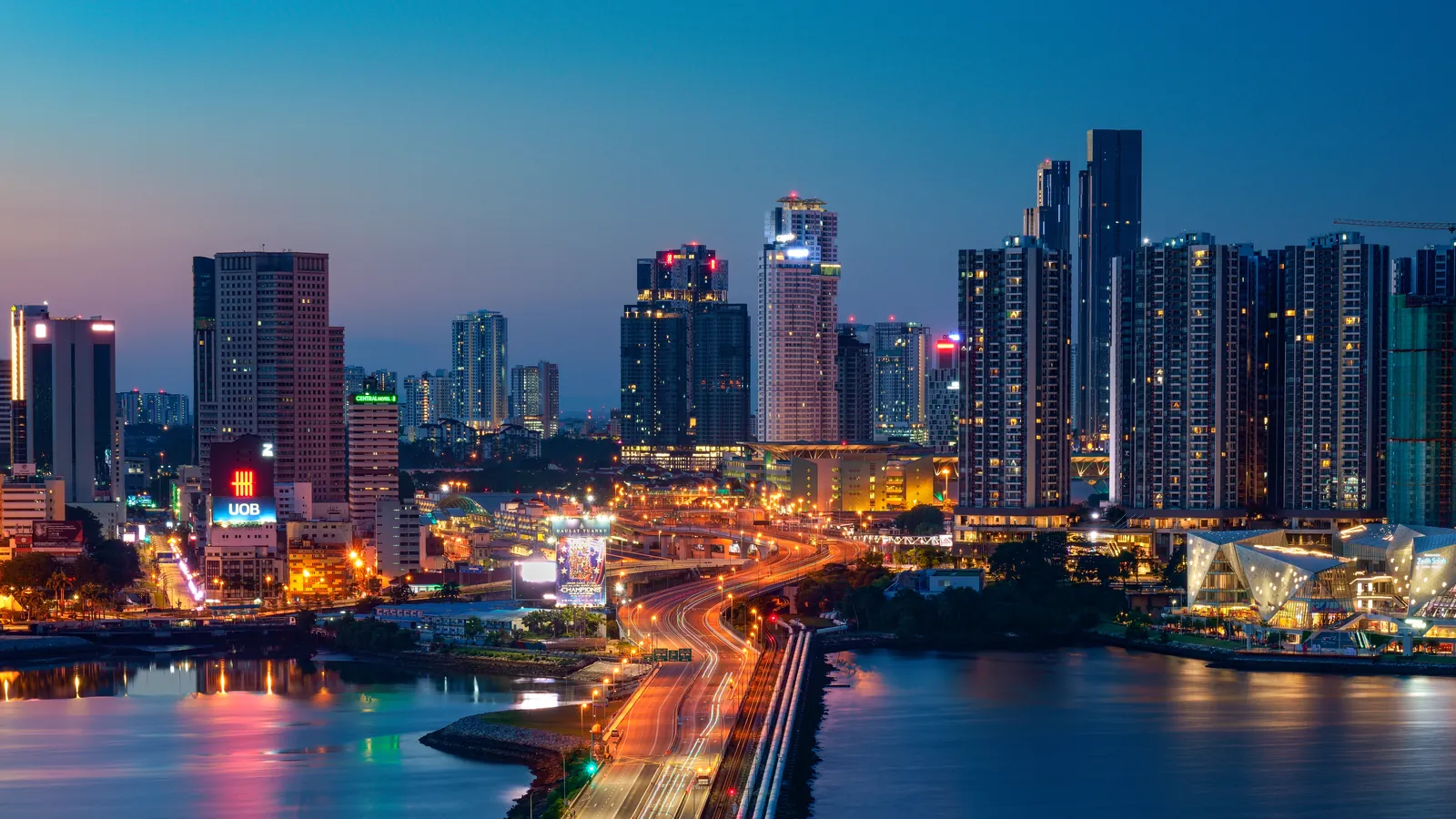 Johor Bahru city skyline lit up at dusk above the Strait of Johor, with the Causeway approach road in the foreground