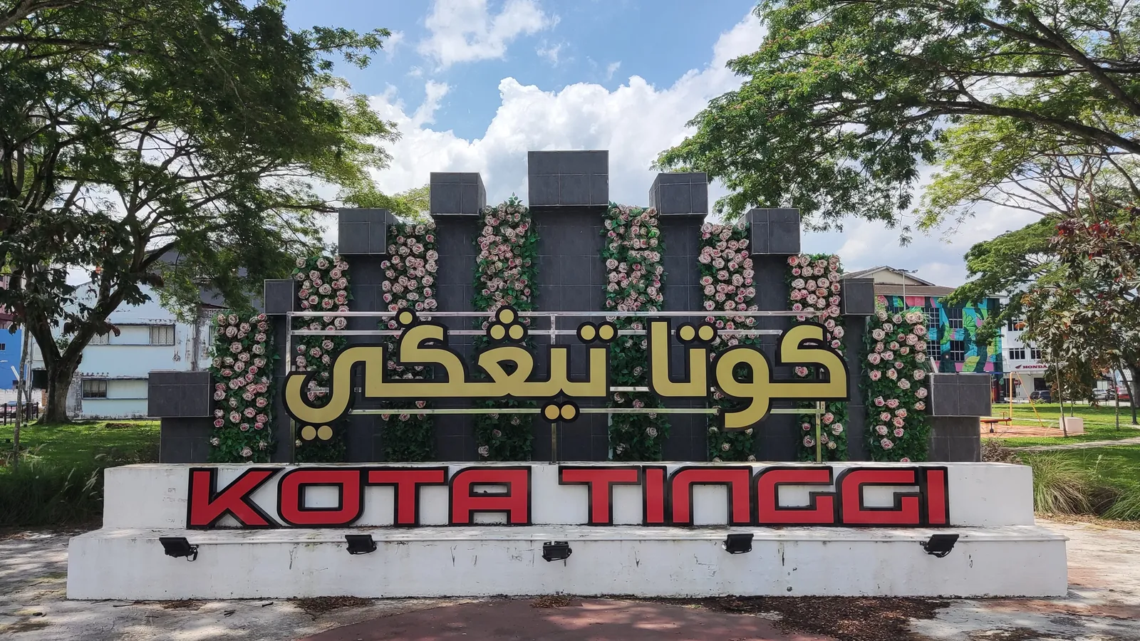 Kota Tinggi town sign in red and Jawi lettering, framed by flowers and shady trees in a riverside park
