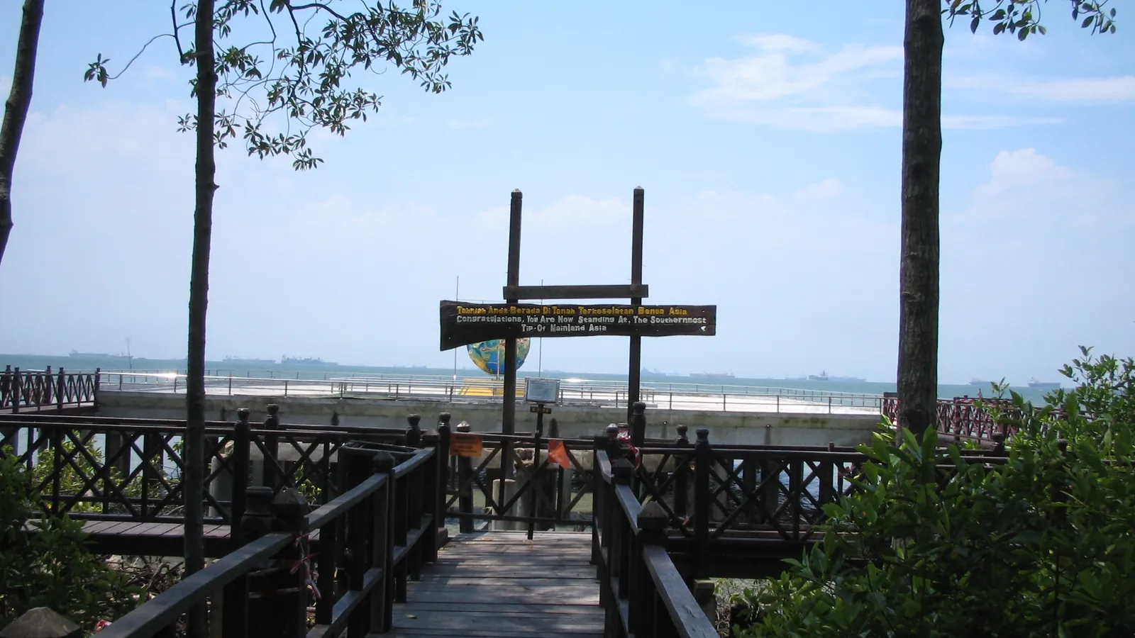 Wooden boardwalk through mangroves leading to the southernmost-point-of-Asia sign at Tanjung Piai, with ships on the horizon