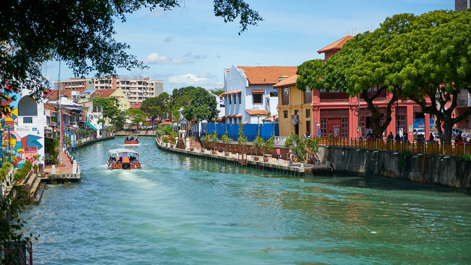 Melaka River lined with colourful heritage shophouses and trees, with a river cruise boat on the water