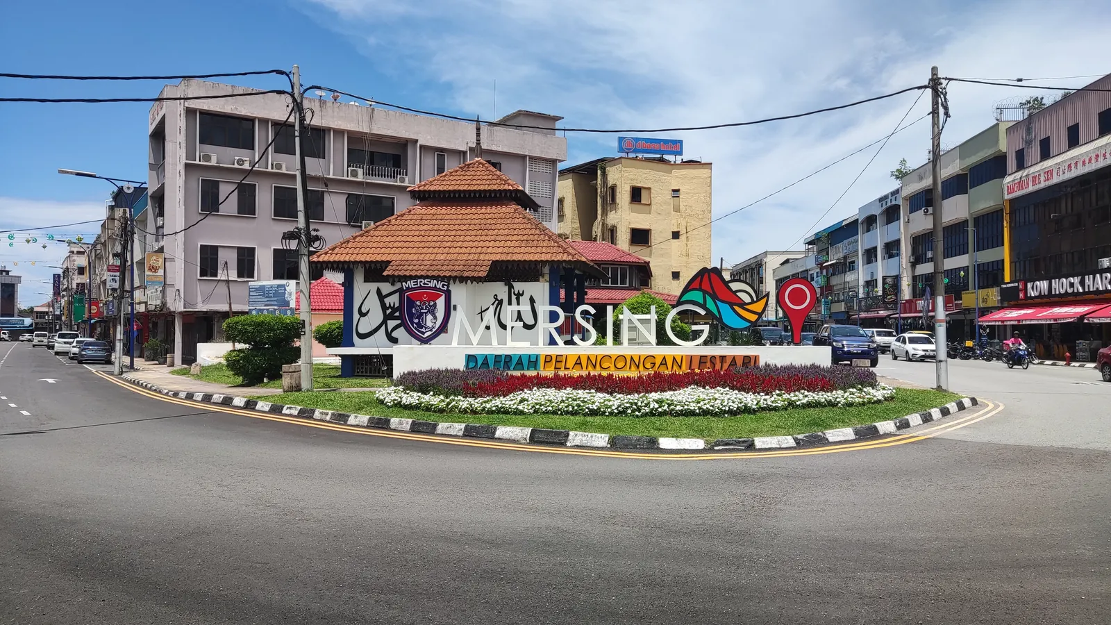 Roundabout in Mersing town, Johor, with a colourful Mersing sign and flower bed in front of shophouses under a blue sky