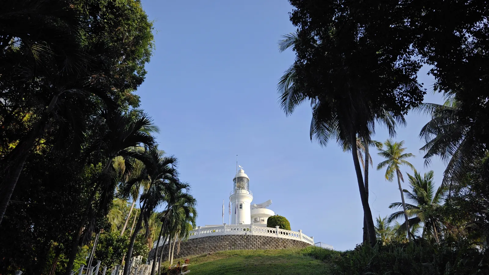 White Cape Rachado Lighthouse on the forested Tanjung Tuan headland near Port Dickson, framed by trees under a blue sky
