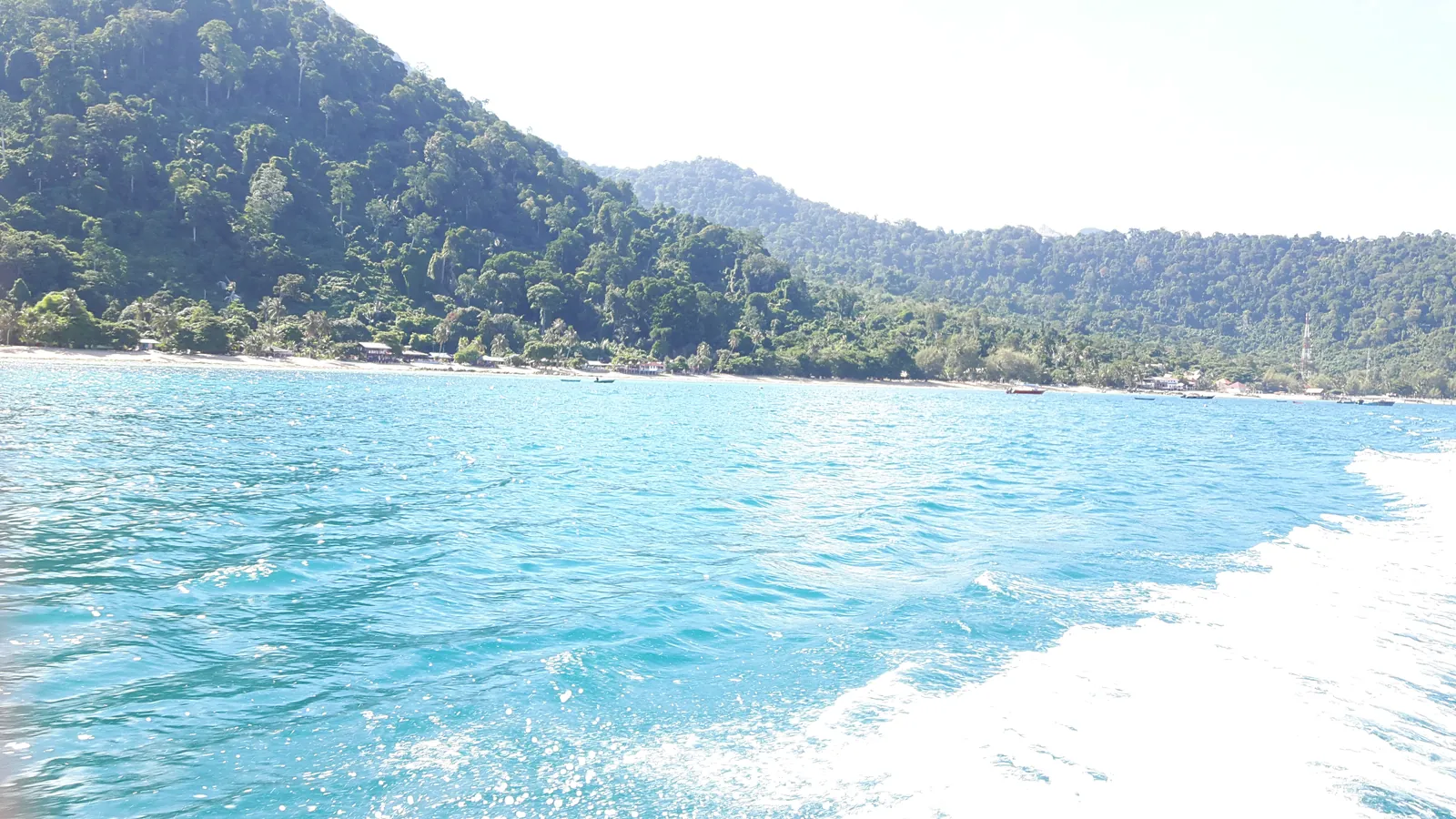 Forested green hills of Tioman Island, Malaysia, seen from a boat over turquoise water with a white wake