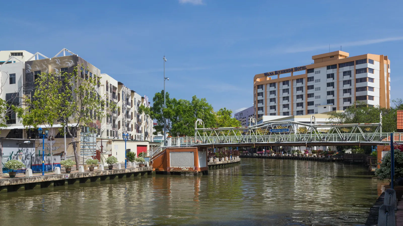 Melaka River with a footbridge, riverside apartments and a hotel tower under a blue sky